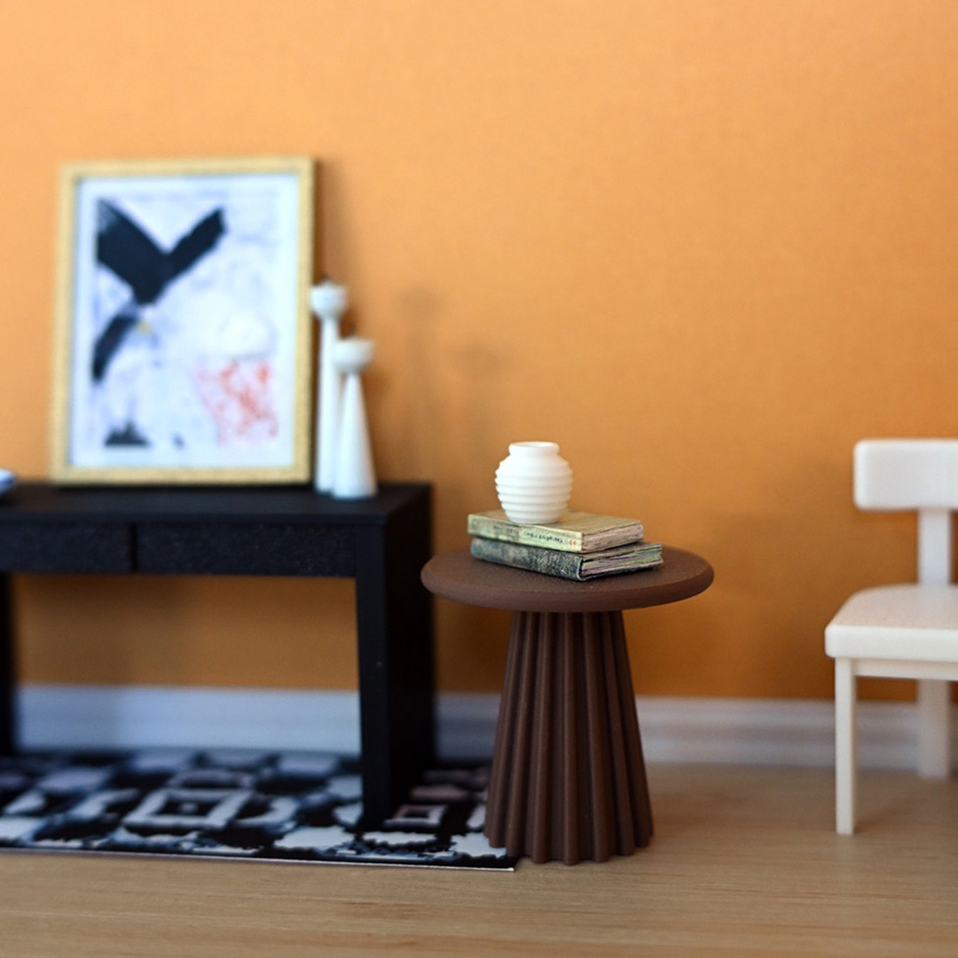 a mini room set-up with the runner black and white rug under a mini 3d printed black desk with painting propped on top beside a chair and table